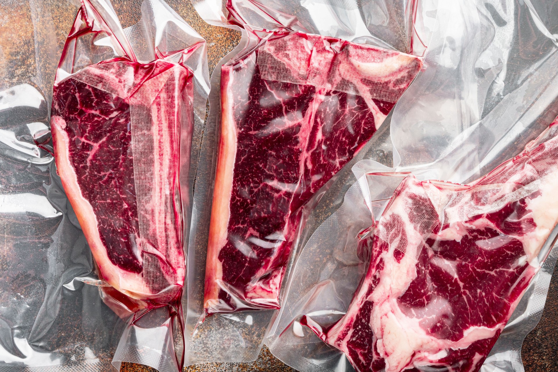 Dry aged steak in a vacuum. Meat products in plastic pack, tomahawk, t bone and club steak cuts, on old dark rustic background, top view flat lay